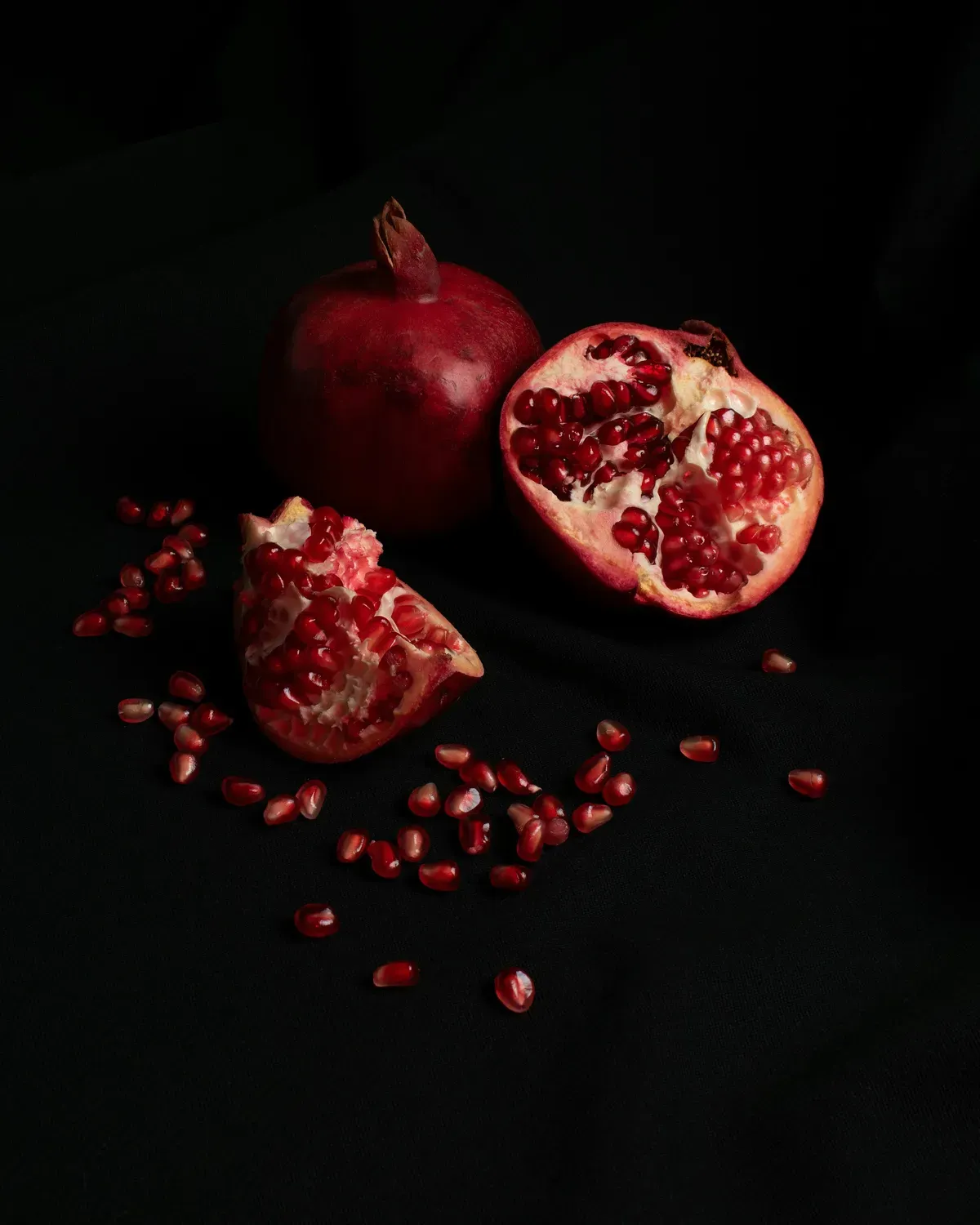 A whole pomegranate and two broken pieces, their red seeds scattered on a dark cloth.